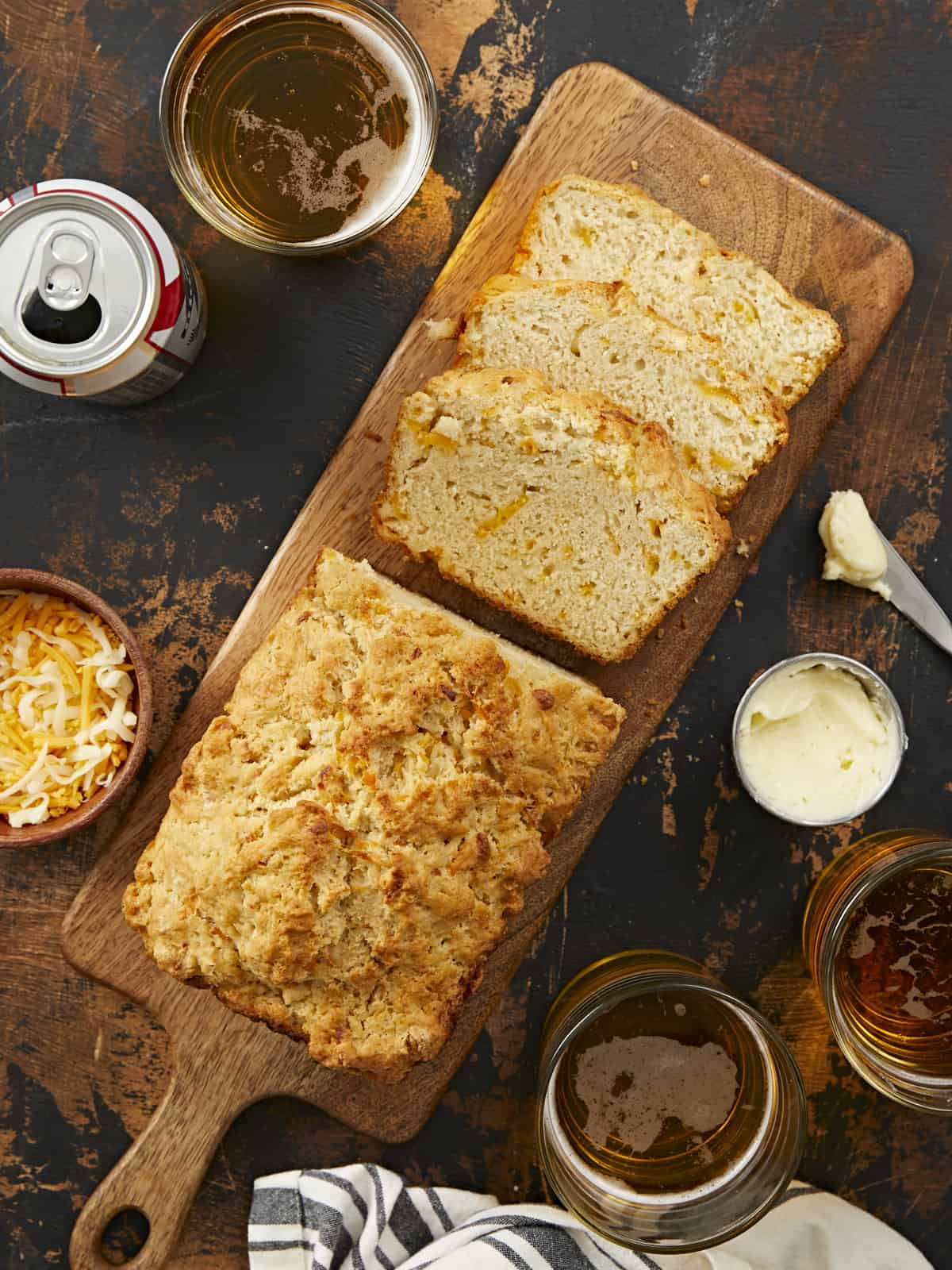 A loaf of beer bread with three slices overlapping on a long wooden cutting board surrounded by glass jars filled with beer, a PBR can, a wooden bowl of shredded cheese and a small metal dish of soft butter and a small butter knife.