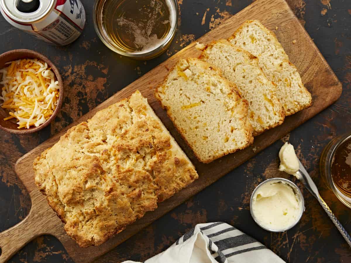 An overhead shot of a half-sliced loaf of beer bread on a long, wooden cutting board surrounded by glass cups of beer, a can of beer, a butter dish and knife, and bowl of cheese, and a grey and white striped dish cloth.