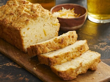 A side shot of a loaf of beer bread on a wooden cutting board with three slices slightly overlapping in the foreground of the image, and behind the cutting board is a small wooden bowl filled with shredded cheese and a glass cup of beer is visible in the upper right corner of the frame.