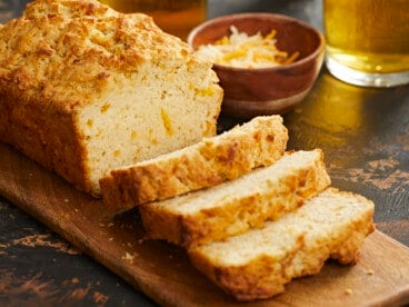 A side shot of a loaf of beer bread on a wooden cutting board with three slices slightly overlapping in the foreground of the image, and behind the cutting board is a small wooden bowl filled with shredded cheese and a glass cup of beer is visible in the upper right corner of the frame.