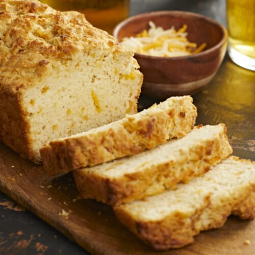 A side shot of a loaf of beer bread on a wooden cutting board with three slices slightly overlapping in the foreground of the image, and behind the cutting board is a small wooden bowl filled with shredded cheese and a glass cup of beer is visible in the upper right corner of the frame.