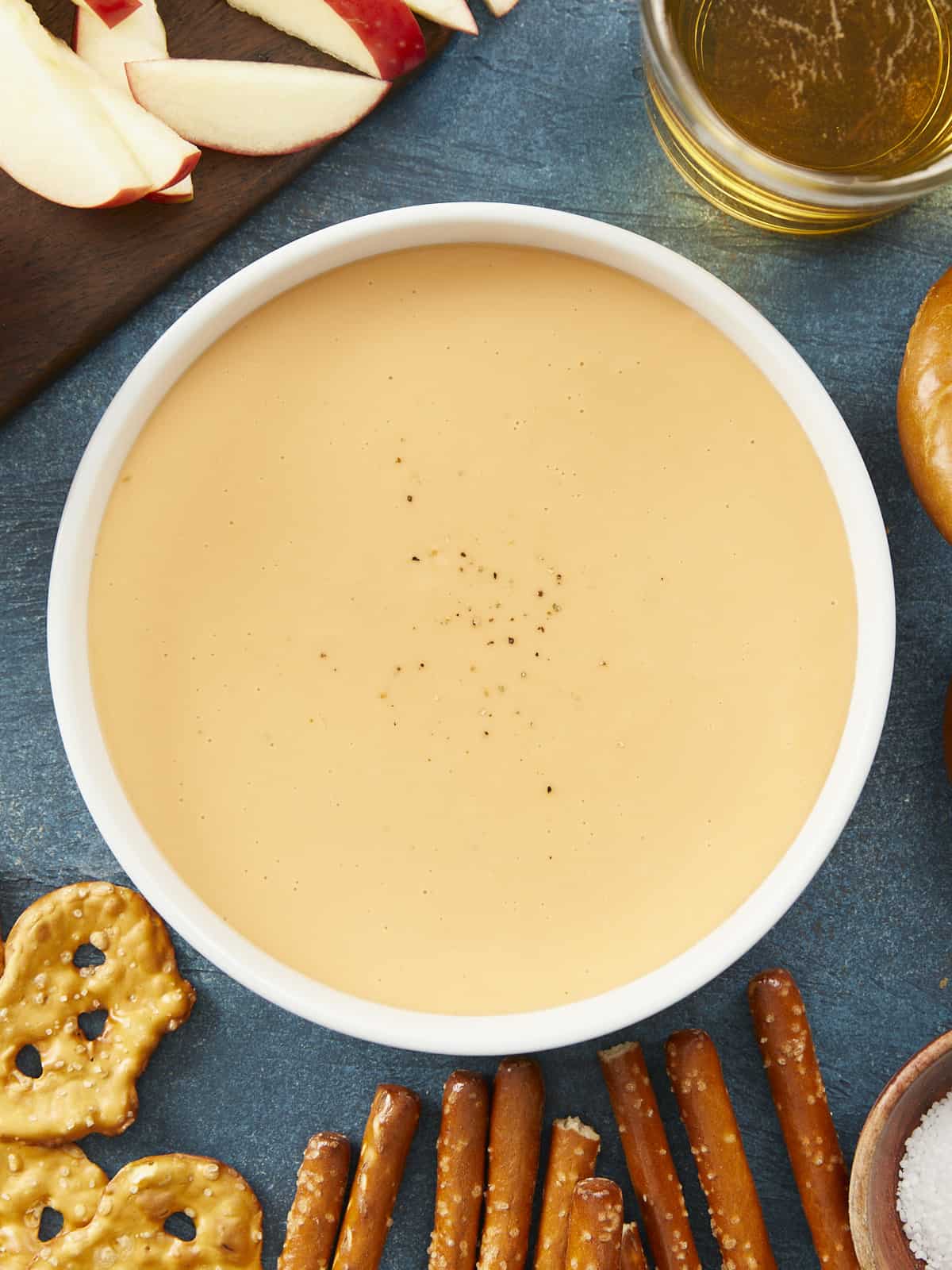 A large white bowl filled with beer cheese dip on a blue background surrounded by different types of pretzels, a wooden cutting board with apple slices and a glass cup of beer.