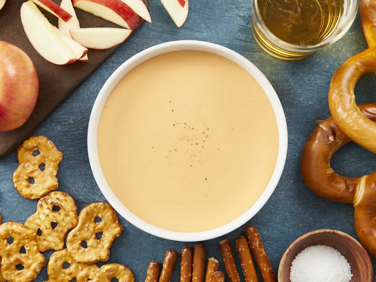 A overhead view of a white bowl of cheese dip sprinkled with black pepper that is surrounded by an assortment of pretzels, a bowl of pretzel salt, a wooden cutting board of apples and a glass cup of beer.