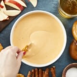 An overhead view of a hand swirling a pretzel crisp into a white bowl of beer cheese dip that's surrounded by an assortment of pretzels, a glass cup of beer, a wooden cutting board topped with apple slices, and a small bowl of pretzel salt.