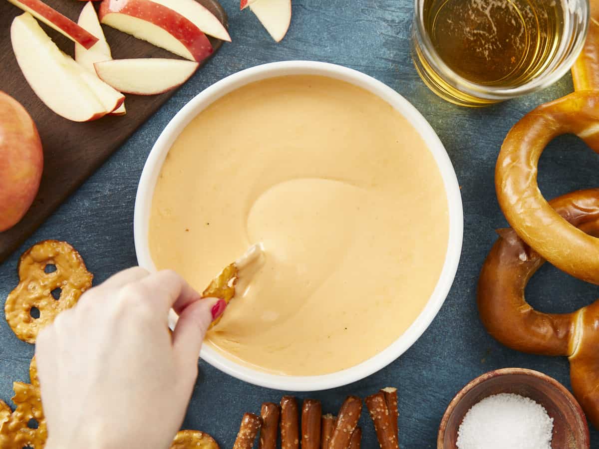 An overhead view of a hand swirling a pretzel crisp into a white bowl of beer cheese dip that's surrounded by an assortment of pretzels, a glass cup of beer, a wooden cutting board topped with apple slices, and a small bowl of pretzel salt.
