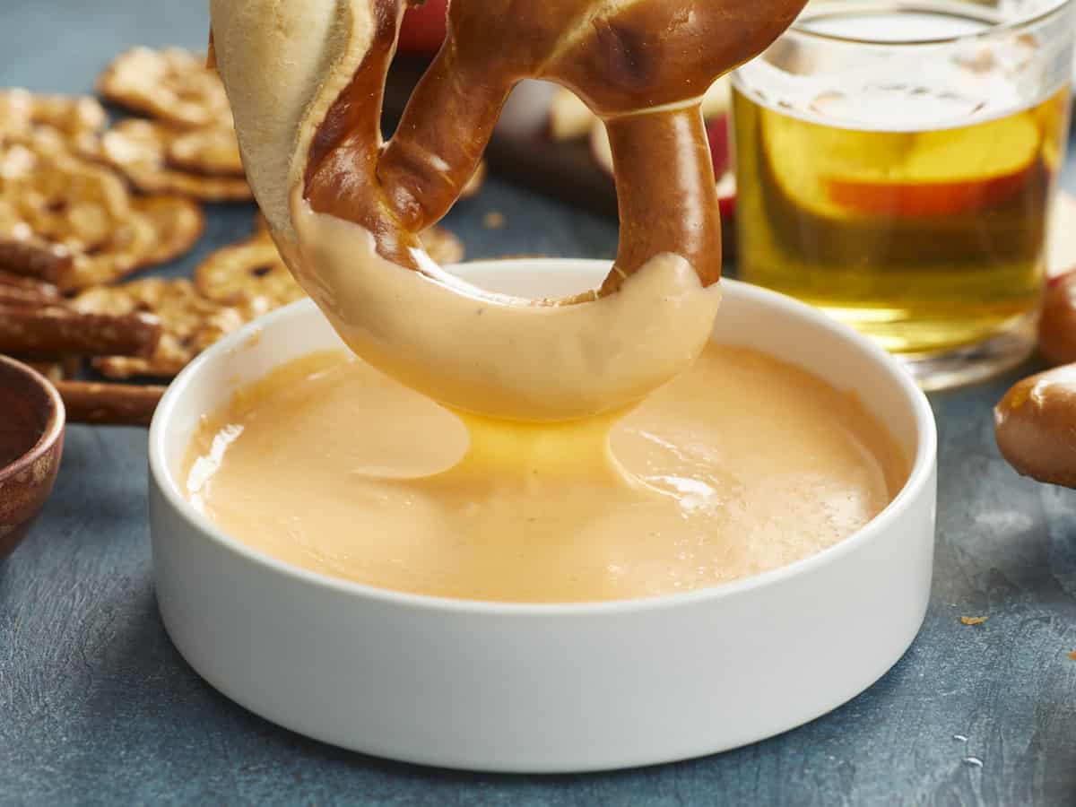 A side view of giant Bavarian pretzel that's just been half-dipped in beer cheese is hovering over a large white bowl filled of the dip and is surrounded by an assortment of other pretzels and a glass cup of beer.