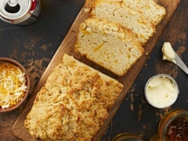 A loaf of beer bread with three slices overlapping on a long wooden cutting board surrounded by glass jars filled with beer, a PBR can, a wooden bowl of shredded cheese and a small metal dish of soft butter and a small butter knife.