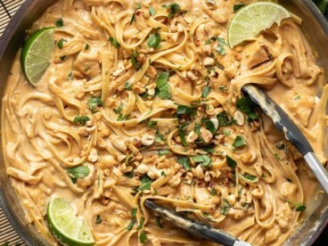 Overhead view of peanut noodles with chicken in a skillet with tongs.