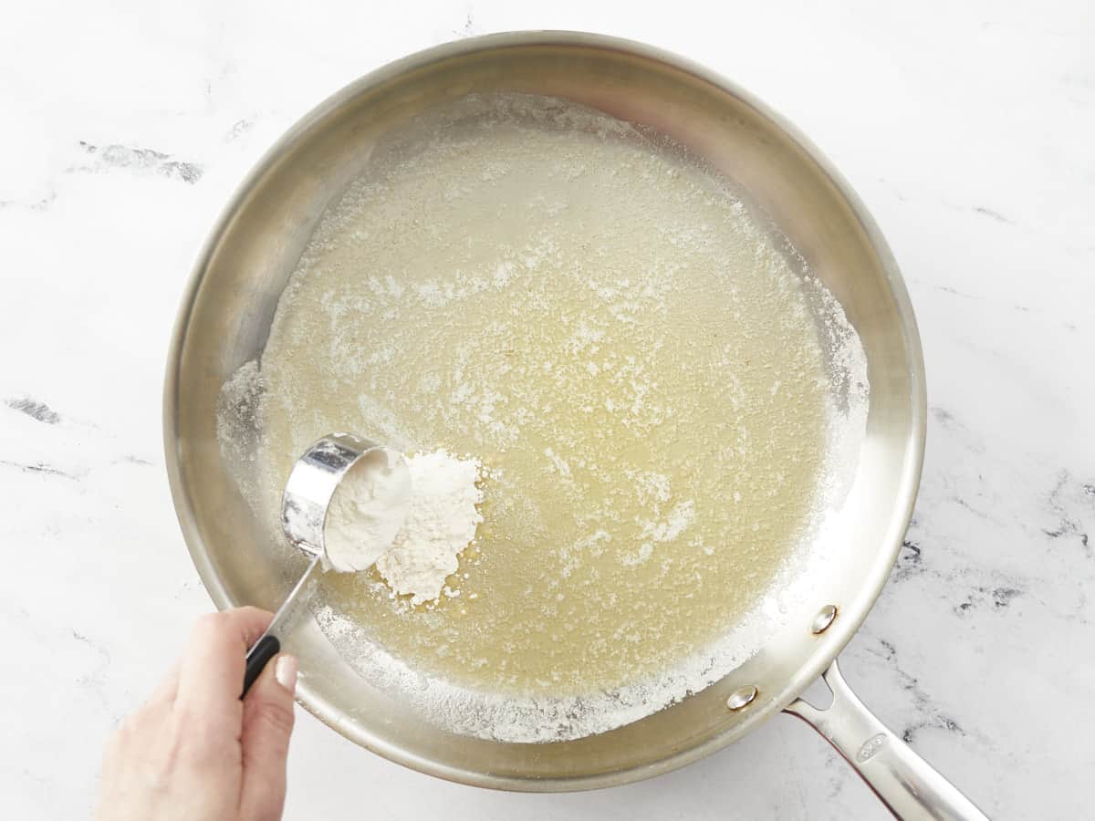 Overhead shot of silver pan with melted butter and a hand adding flour with a silver scoop.