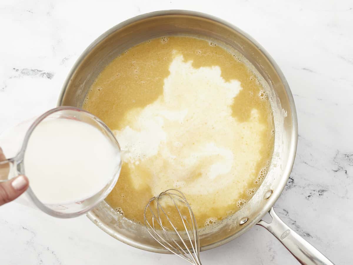 Overhead shot of cream being in a measuring cup being added to sauce in a silver pan with a whisk in it.