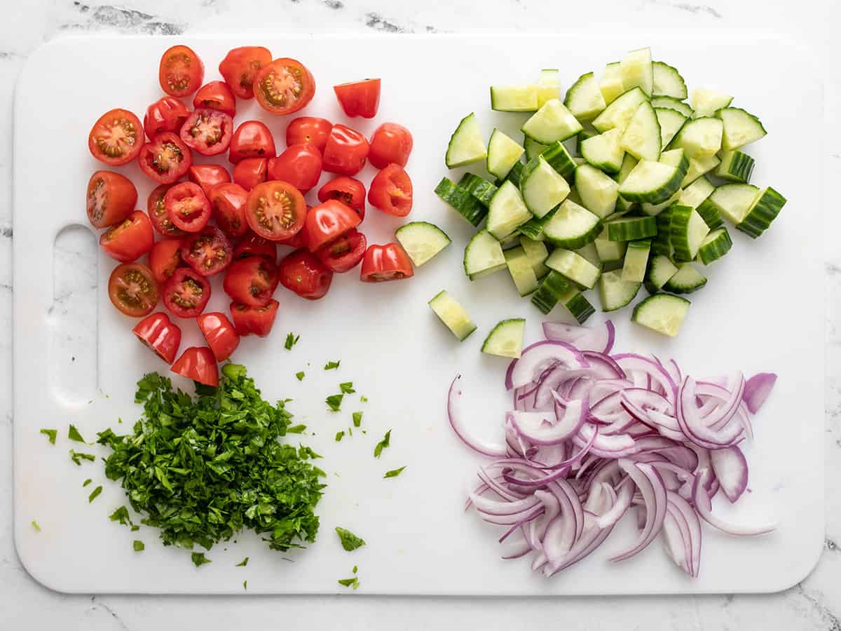 Tomates preparados, pepinos, cebollas y perejil en una tabla para cortar.