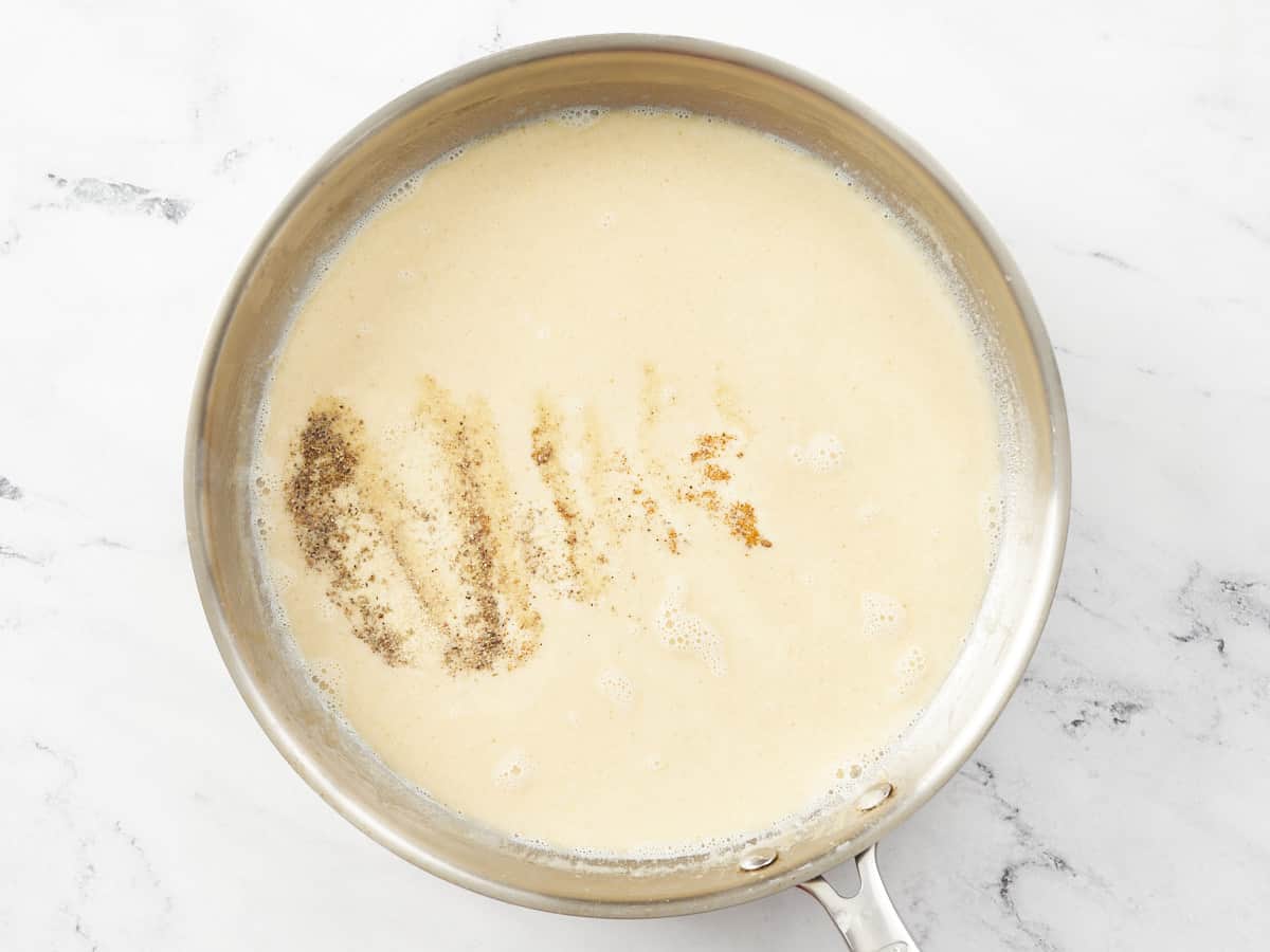 Overhead shot of a cream sauce in a silver pan with with spices being added to it.