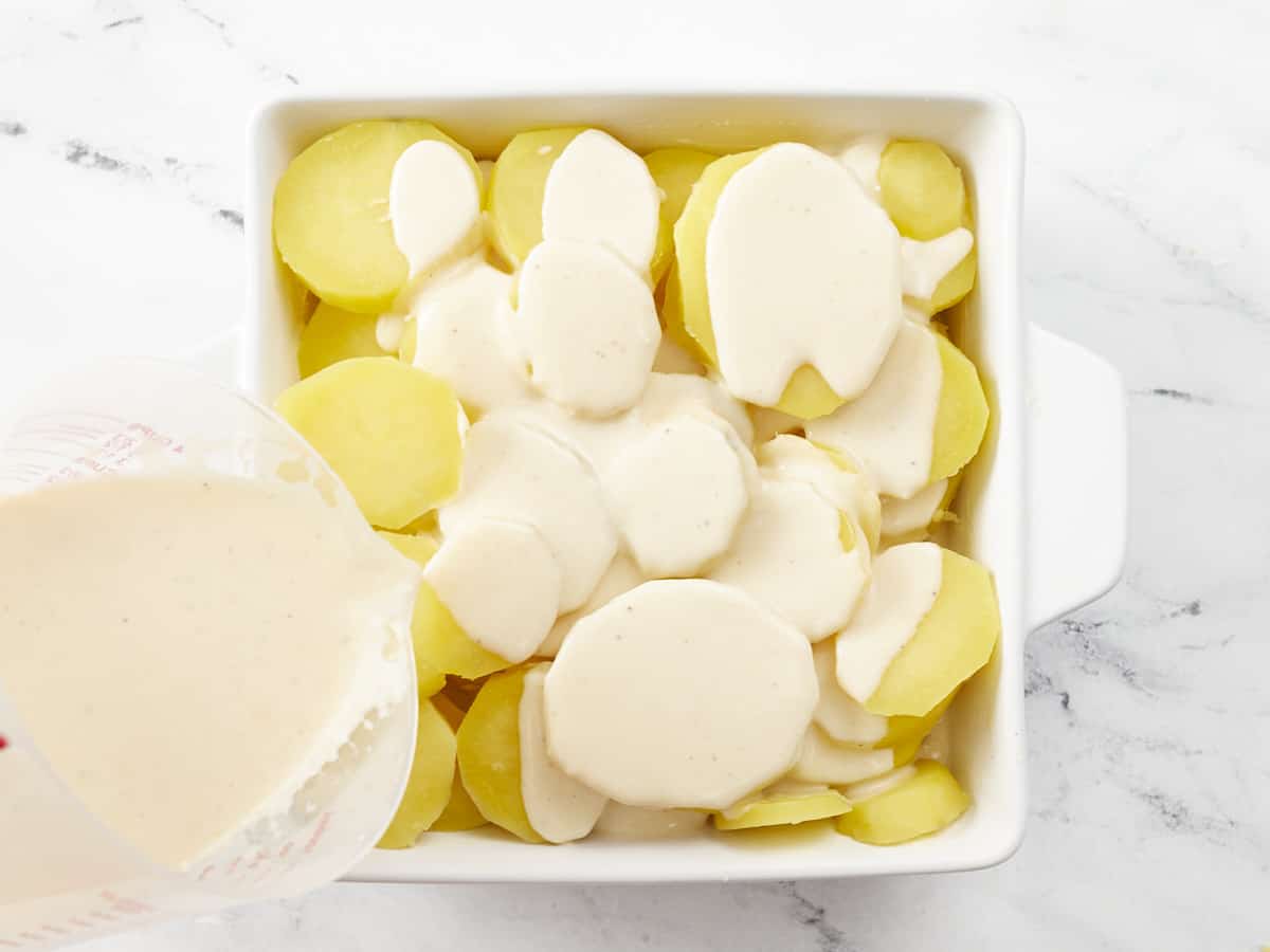 Overhead shot of raw scalloped potatoes in a white casserole dish with cream sauce being poured into it.