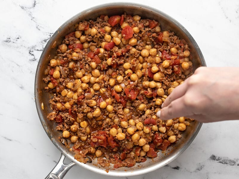 Overhead shot of hand sprinkling red pepper flakes into finished ground beef, tomato, and garbanzo bean mixture.