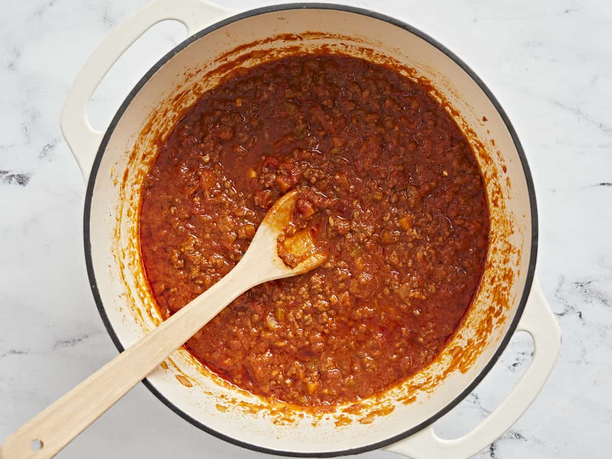 Overhead shot of bolognese in a beige Dutch oven with a wood spoon in it.
