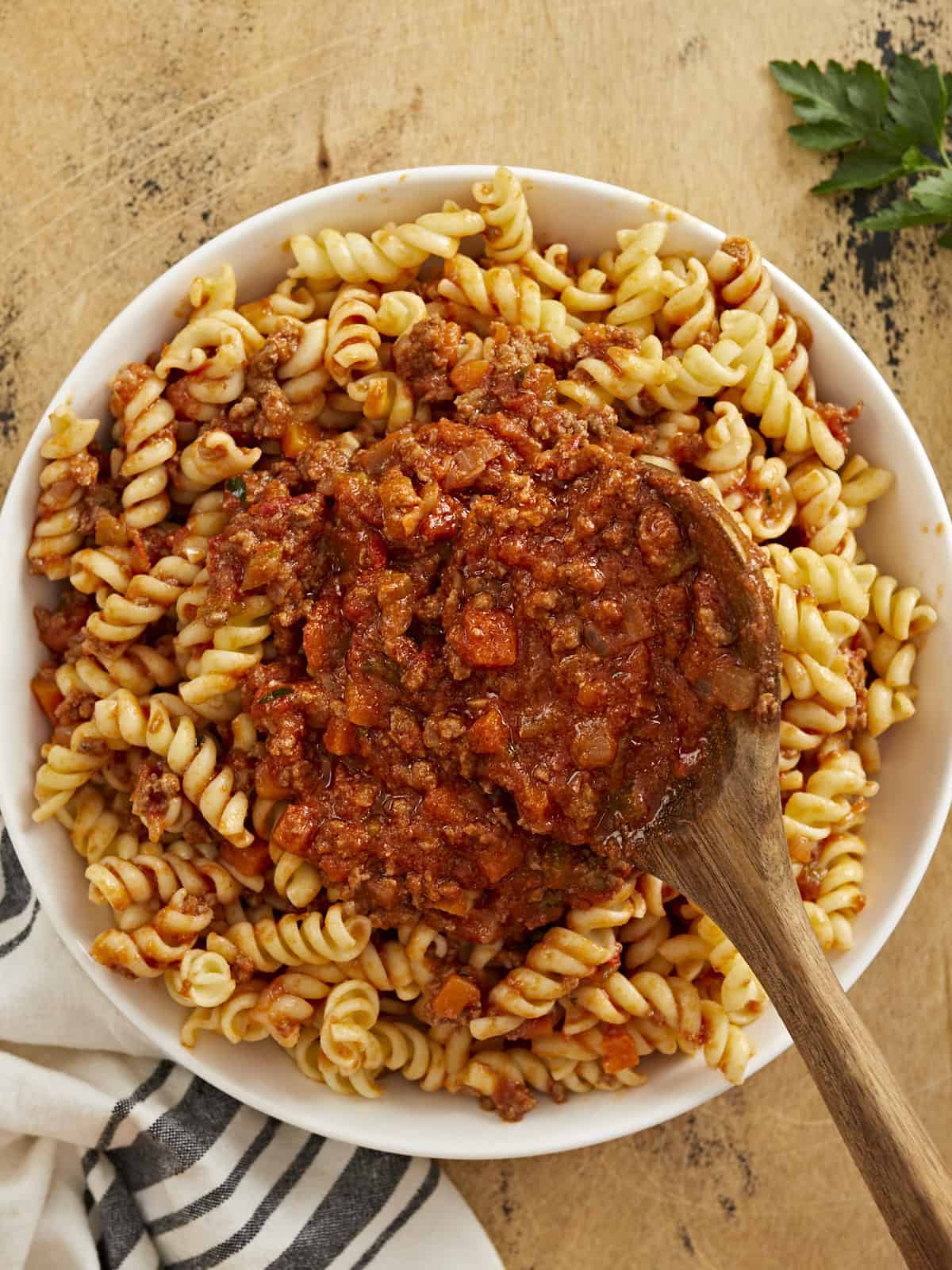 Overhead shot of bolognese served over rotini in a white bowl with a wood spoon in it.