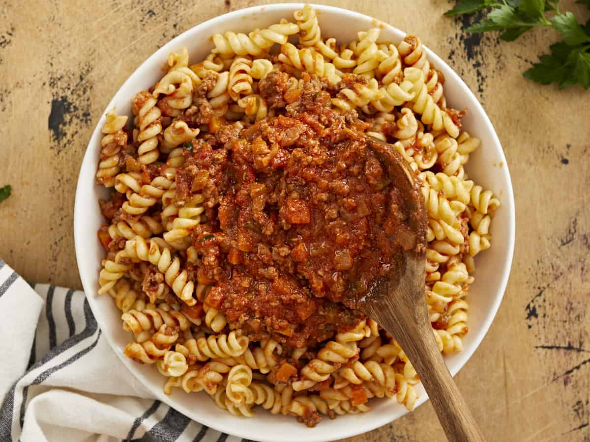 Overhead shot of bolognese served over rotini in a white bowl with a wood spoon in it.
