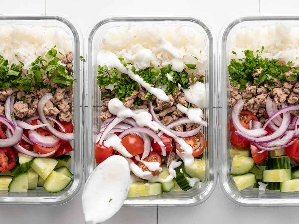 Three meal prep turkey bowls lined up with yogurt sauce being drizzled over the middle one.