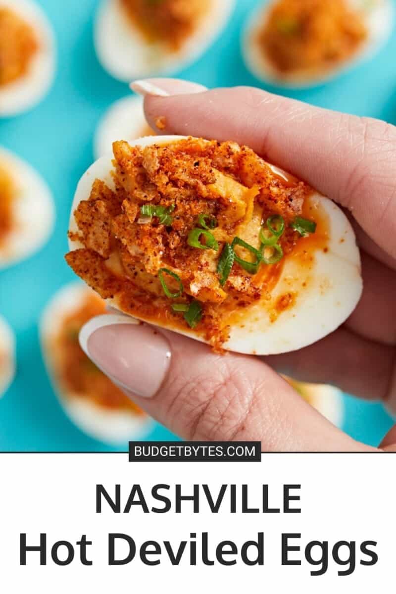 Close up of hand holding a Nashville Hot Deviled Egg with other eggs in the background on an aqua backdrop.