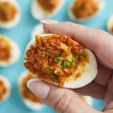 Close up of hand holding a Nashville Hot Deviled Egg with other eggs in the background on an aqua backdrop.