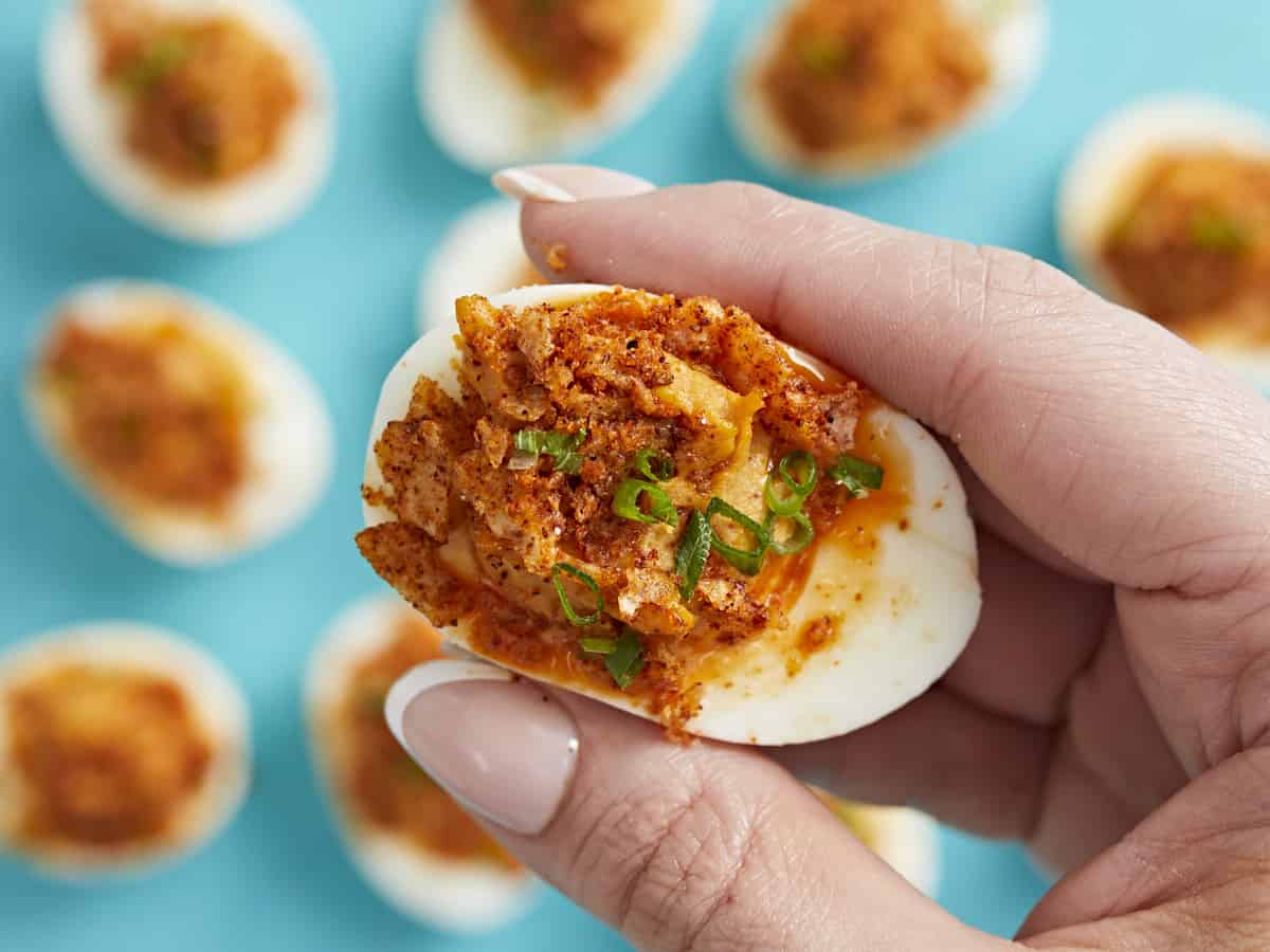 Close up of hand holding a Nashville Hot Deviled Egg with other eggs in the background on an aqua backdrop.