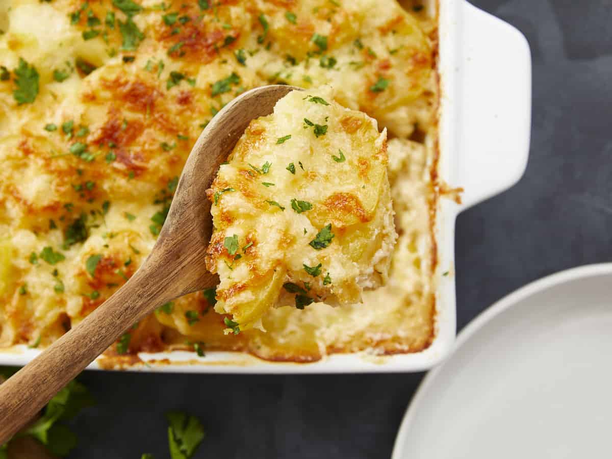 Overhead shot of scalloped potatoes in a white casserole dish with a wooden spoon over it holding a spoonful of potatoes.