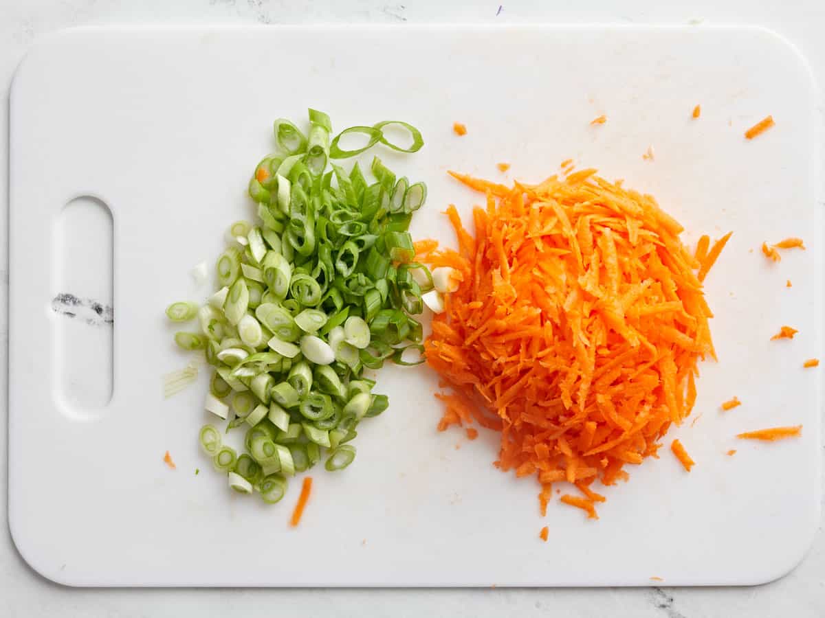 Sliced green onion and shredded carrot on a cutting board. 