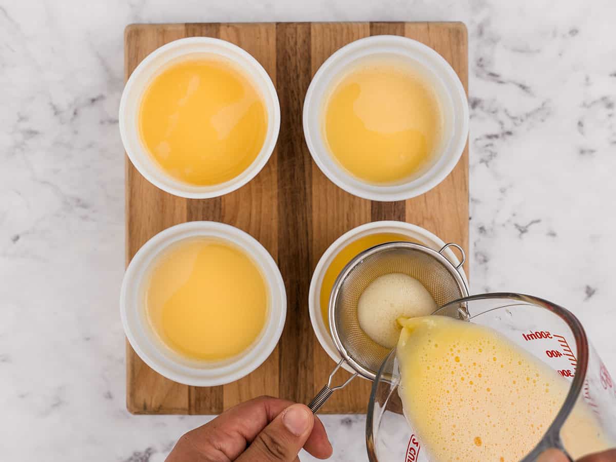 Egg mixture being poured through a fine mesh sieve into white ramekins.