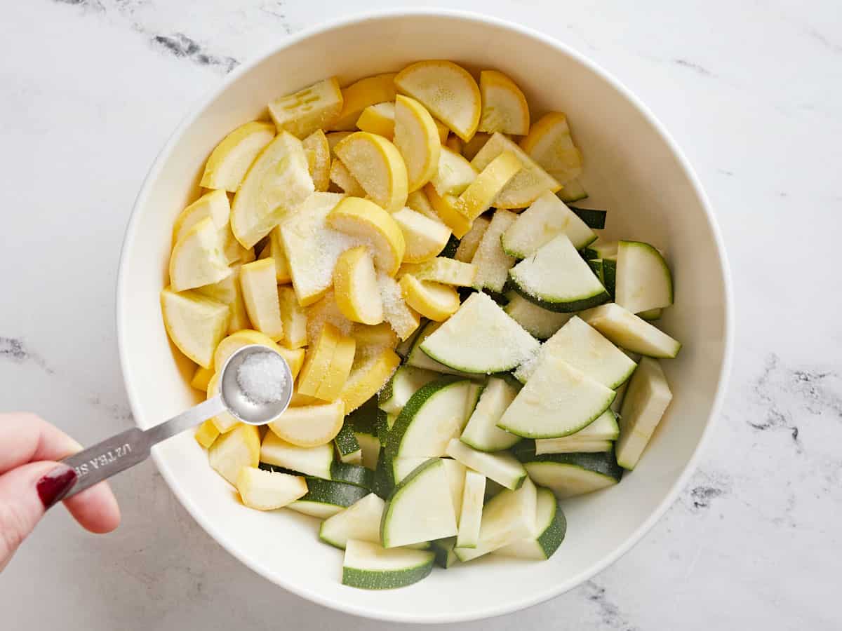 Sliced zucchini and squash in a bowl with salt being sprinkled over top. 