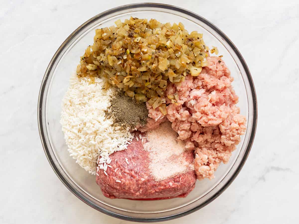 Overhead view of large glass bowl with ground pork, ground beef, uncooked white rice, sautéed onion, garlic and seasoning.