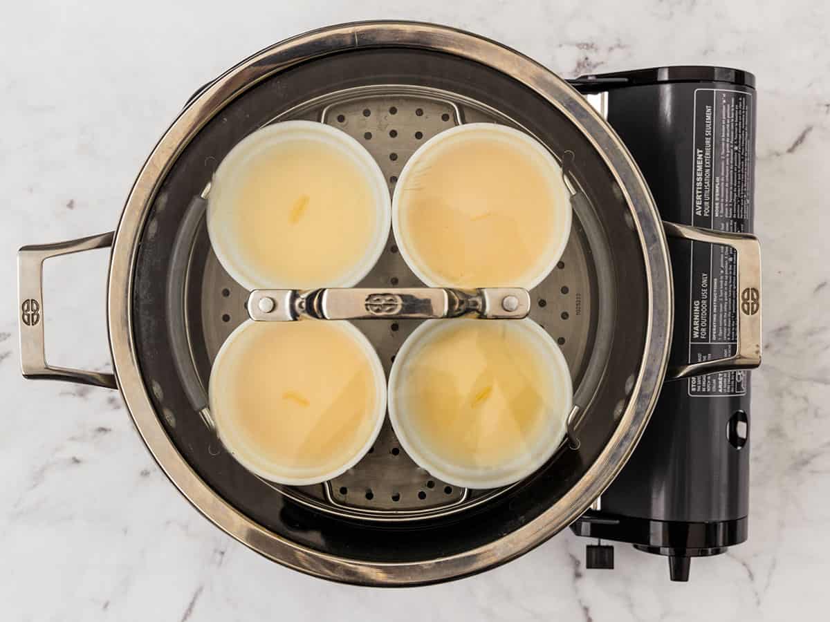 Ramekins filled with egg mixture sitting on top of a steamer basket over a pot of boiling water and covered with a glass lid.