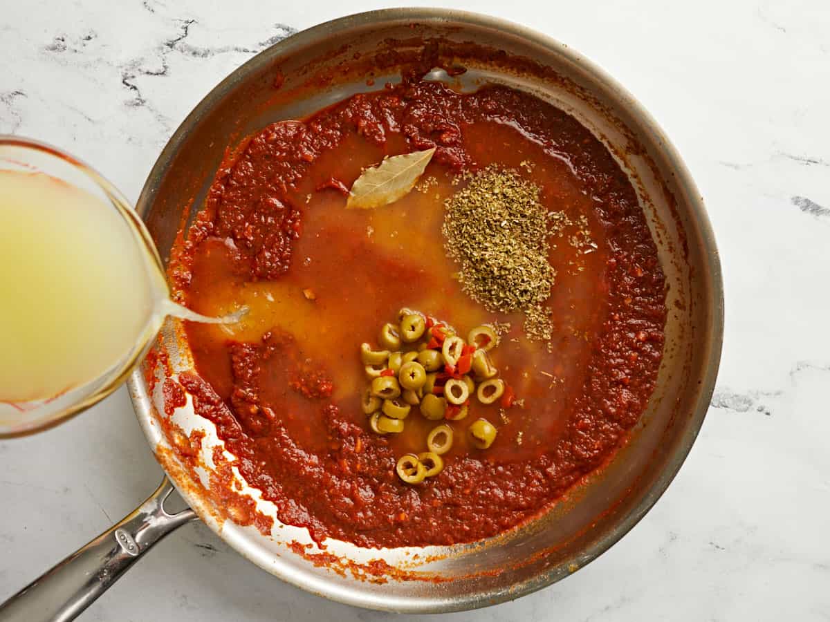 Olives and herbs added to the skillet and broth being poured in the side.