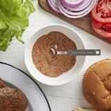 A bowl of burger seasoning surrounded by cooked burger patties and toppings.