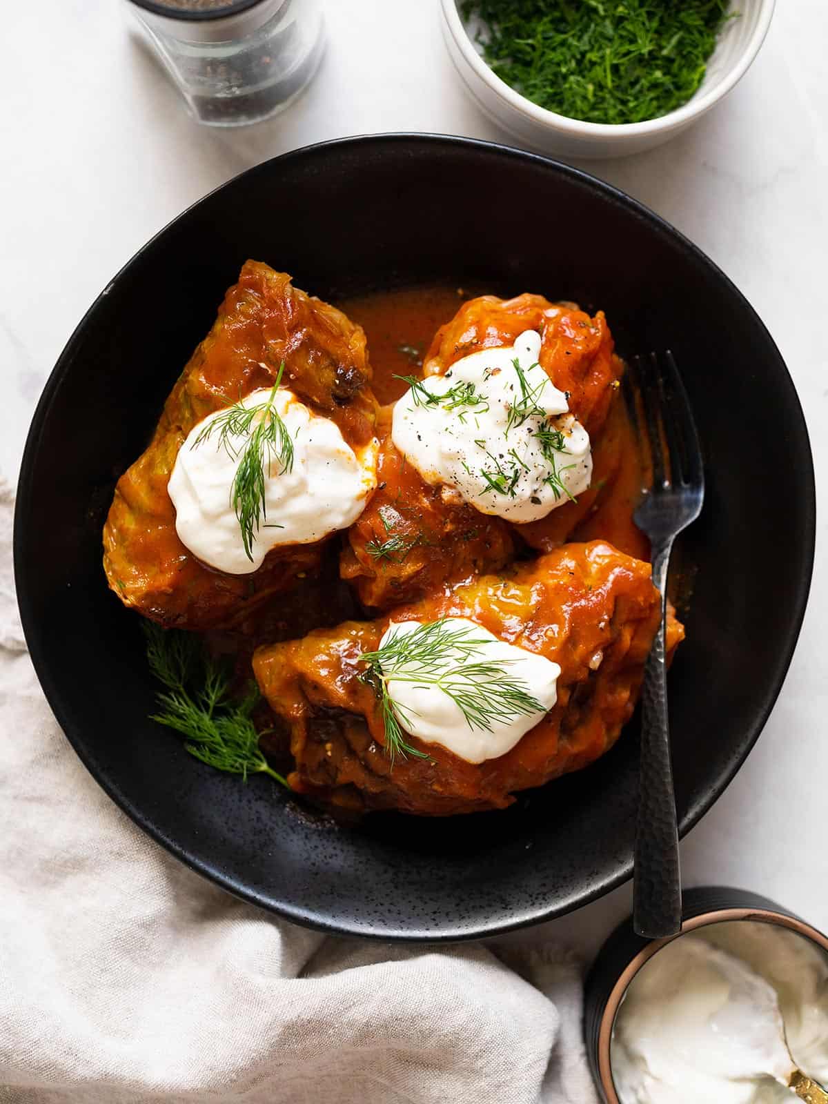 Overhead view of cooked cabbage rolls with tomato sauce on a black serving plate topped with sour cream and fresh dill.