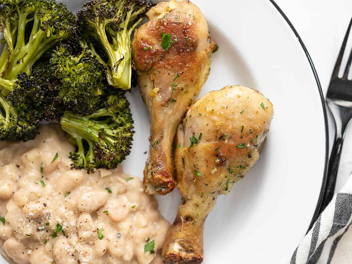 Close up view of two ranch chicken drumsticks on a plate with Parmesan beans and roasted broccoli.