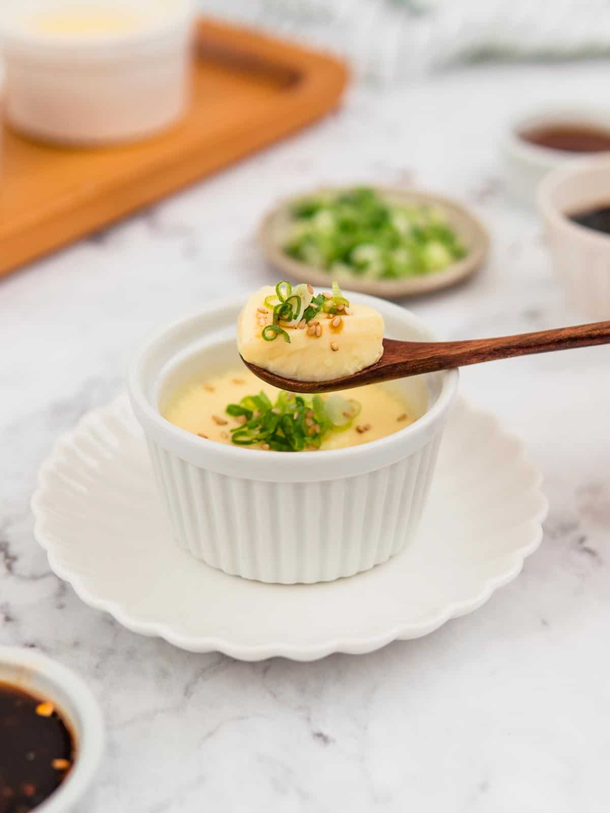 Steamed eggs in a white ramekin garnished with green onions and a spoon lifting some eggs out of the ramekin.