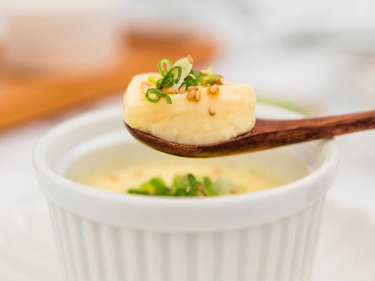 Steamed eggs in a white ramekin garnished with green onions and a spoon lifting some eggs out of the ramekin.