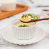 Steamed eggs in a white ramekin garnished with green onions and a spoon lifting some eggs out of the ramekin.