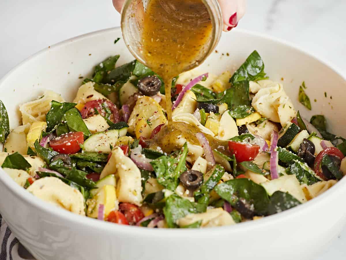 Side shot of tortellini salad in a white bowl with dressing being poured in.