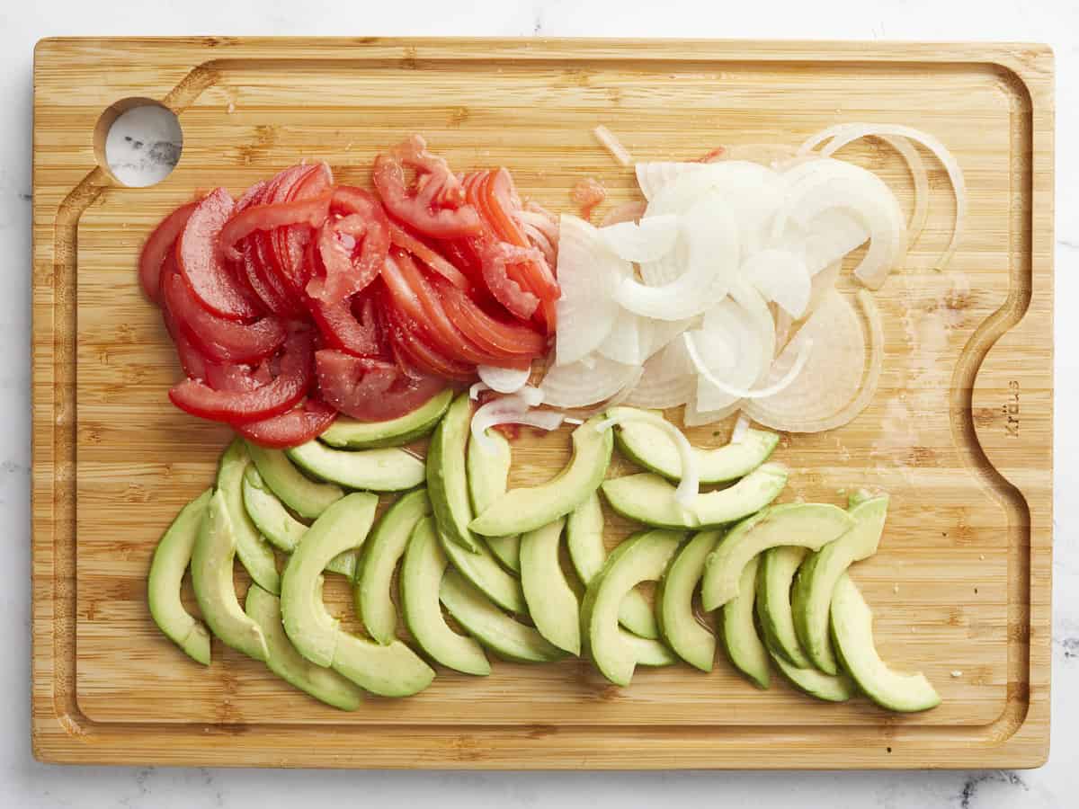 Overhead shot of slice avocados, tomatoes, ad onions on a wood cutting board.