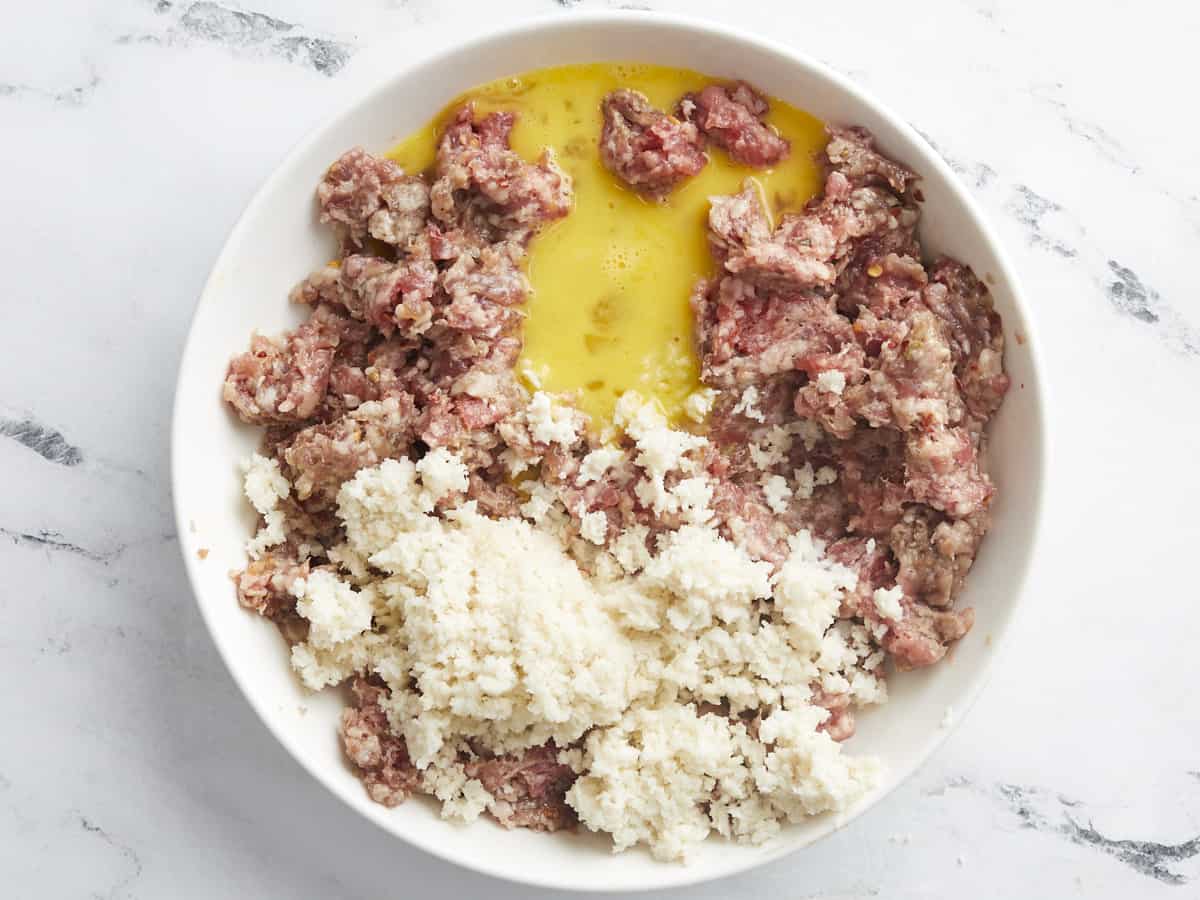 Overhead shot of ground Italian Sausage, breadcrumbs, and egg in a white bowl.