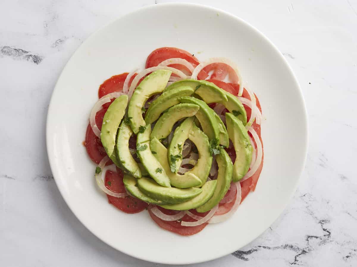 Overhead shot of the first layer of tomatoes, avocados, and onion in Ensalada de Aguacate on a white plate.