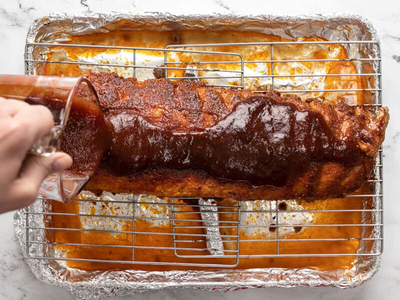 Adding BBQ sauce to baked ribs on a rack.