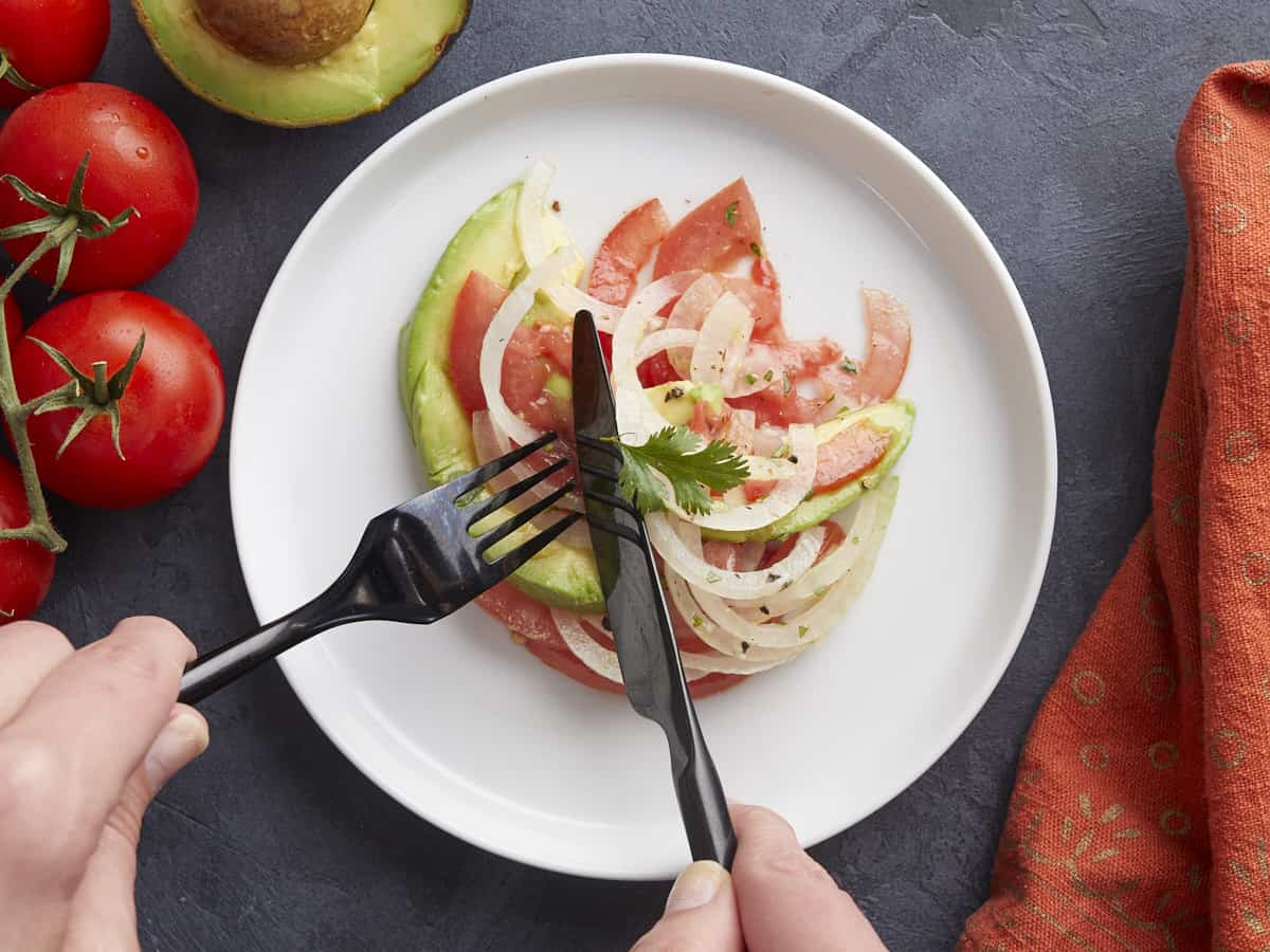 Overhead shot of Ensalada de Aguacate on a white plate with a black fork and knife cutting into it.