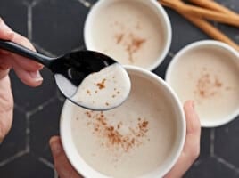 Overhead shot of three white bowls of Maizena with cinnamon sprinkled on top with one bowl one the forground being held by a hand and with a black spoon being dipped into it.