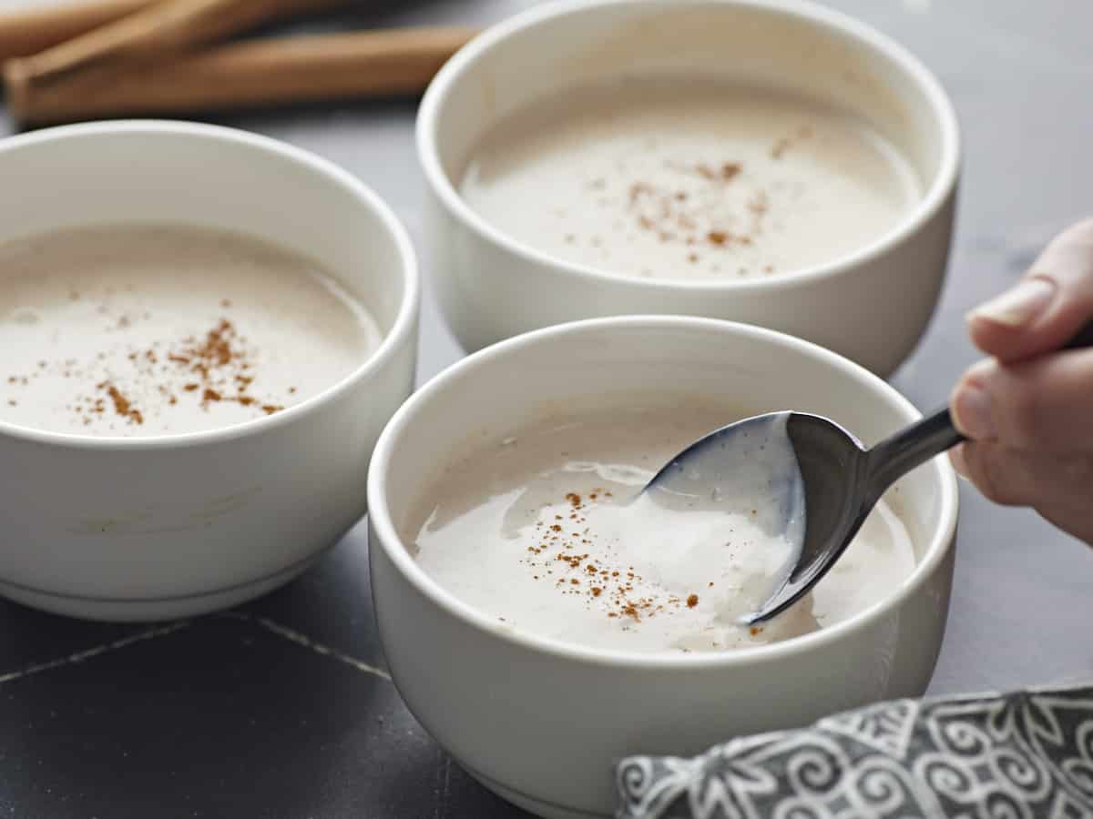 Side shot of three white bowls of Maizena with cinnamon sprinkled on top with one bowl in the foreground being held by a hand and with a black spoon being dipped into it.