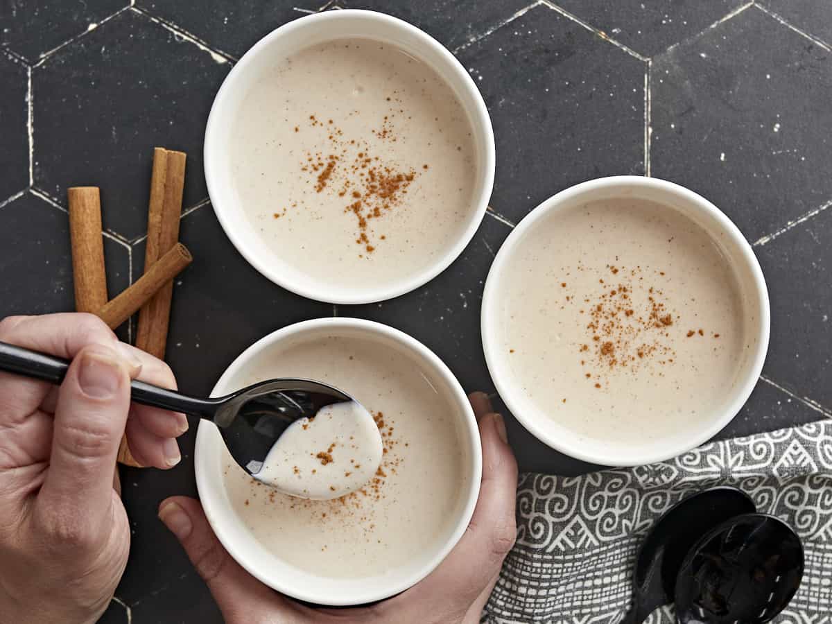 Overhead shot of three white bowls of Maizena with cinnamon sprinkled on top with one bowl in the foreground being held by a hand and with a black spoon being dipped into it.