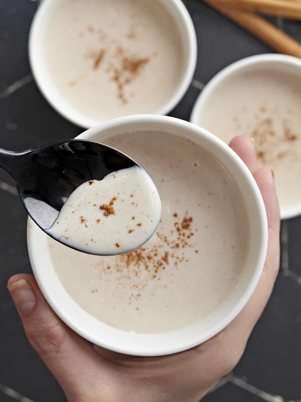 Overhead shot of three white bowls of Maizena with cinnamon sprinkled on top with one bowl in the foreground being held by a hand and with a black spoon being dipped into it.