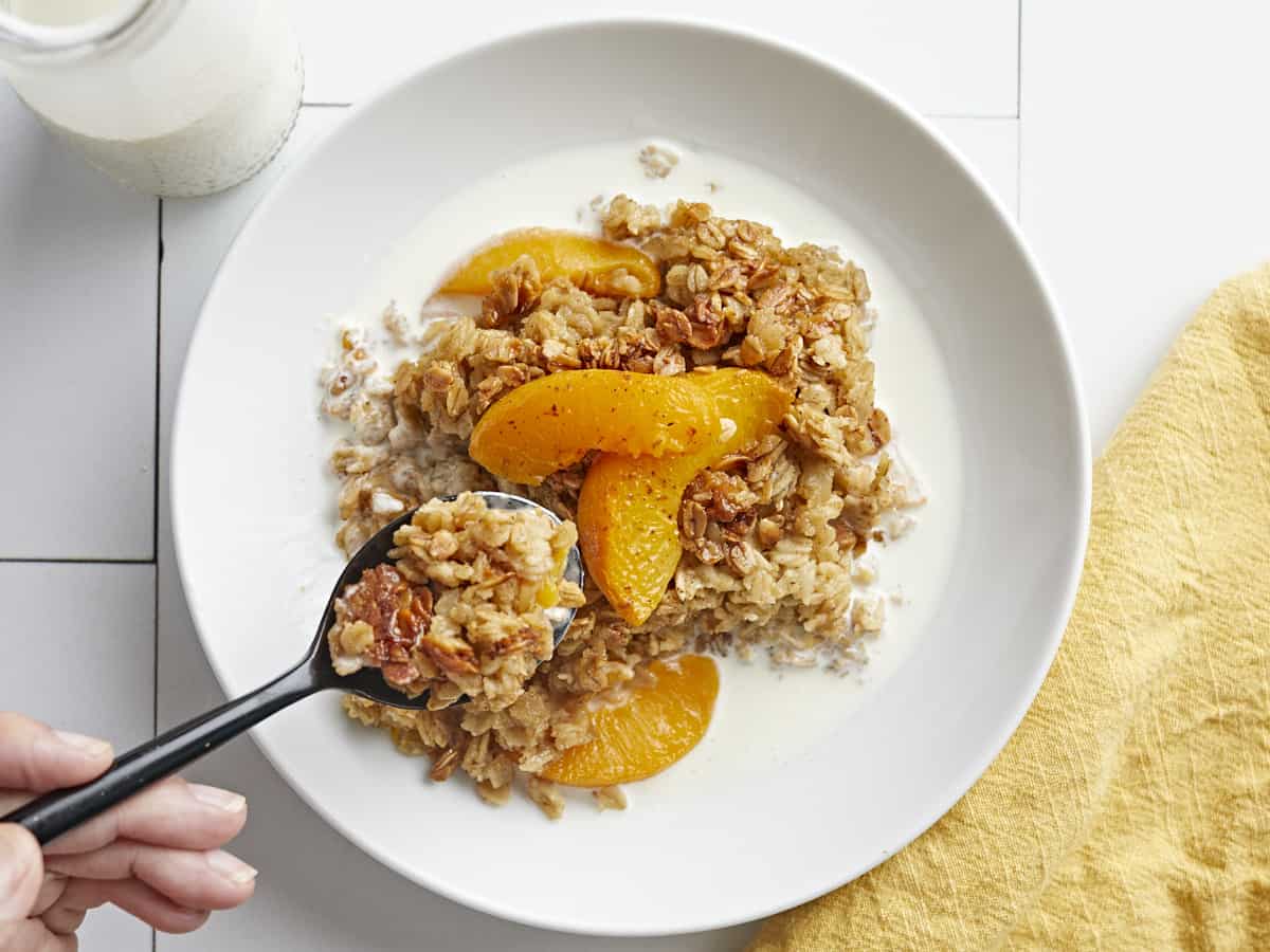 Overhead shot of Peach Baked Oatmeal in a white bowl with cream in it with a spoon lifting a portion out of it.