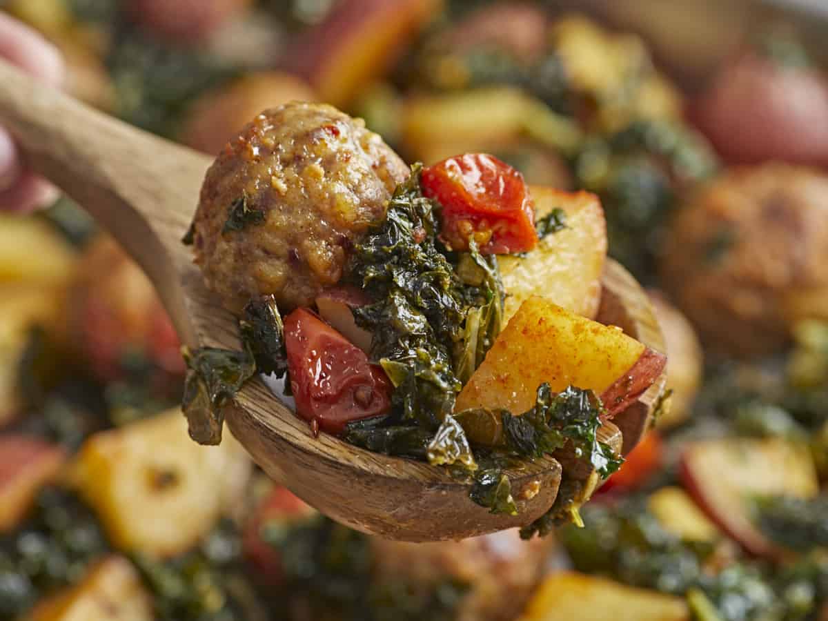 Side shot close up of a wood spoon holding a portion of Sausage Meatball and Kale Sheet Pan Meal.